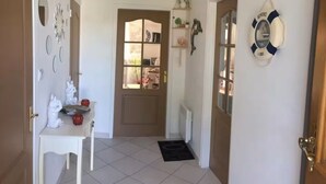 A hallway with a white tiled floor, a white table with decorative items, a door with a glass panel, and a life preserver hanging on the wall. - Maison Sainte Cécile near beach and shops, pets welcome (Camiers)