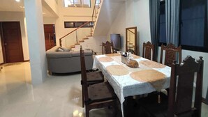A dining area with a table set for four, surrounded by wooden chairs. A staircase leads to an upper level, and a sofa is visible in the background. - 3 Bedroom Exclusive Gated Home in Manila (Las Piñas)