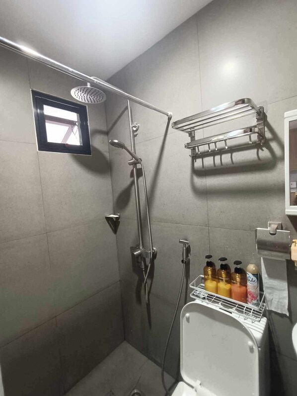 Zona de ducha con cabezal de ducha de mano, toallero adherido a la pared y una ventana pequeña.