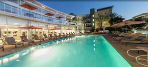 Zona de piscina con tumbonas y sombrillas, un edificio con balcones y un cielo despejado.