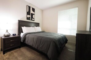 A bedroom with a bed, a nightstand, a lamp, a digital clock, a window with blinds, and a dark-colored dresser.