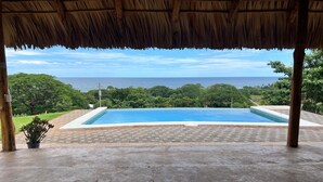 A pool area with a view of the ocean, surrounded by greenery and a thatched roof structure.