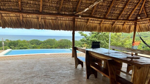 A wooden table and bench under a thatched roof, overlooking a swimming pool and lush greenery.
