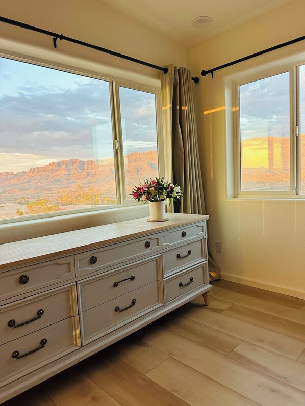 Quarto com uma janela grande, penteadeira branca com várias gavetas e um vaso com flores em cima.