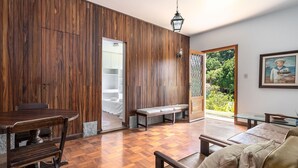 A spacious room with a wooden floor, a dining table with chairs, a sofa, and a bench. There is a door leading to a balcony with a view of greenery.