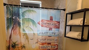 A bathroom with a shower curtain featuring motivational phrases, a toilet, a sink, and a shelf with towels. - Lovely 2-bedroom house with AC in vibrant Miami (Miami)