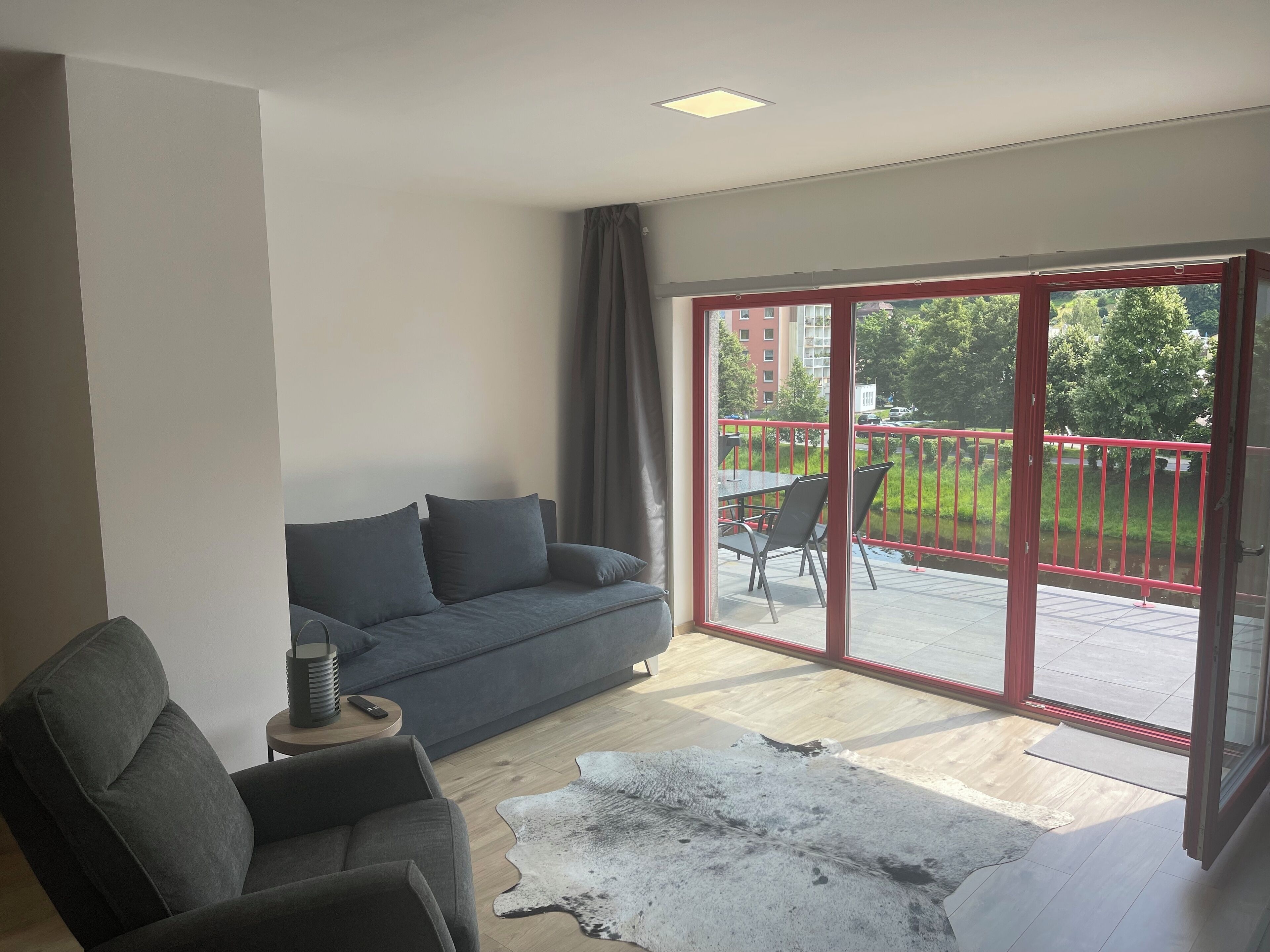 A modern living room with a grey sofa, a small round table with a lamp, and a large sliding glass door leading to an outdoor area with a red railing and greenery.