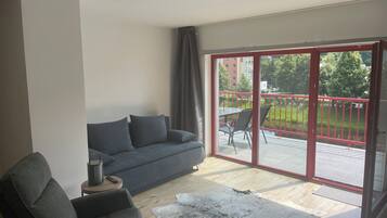 A modern living room with a grey sofa, a small round table with a lamp, and a large sliding glass door leading to an outdoor area with a red railing and greenery.
