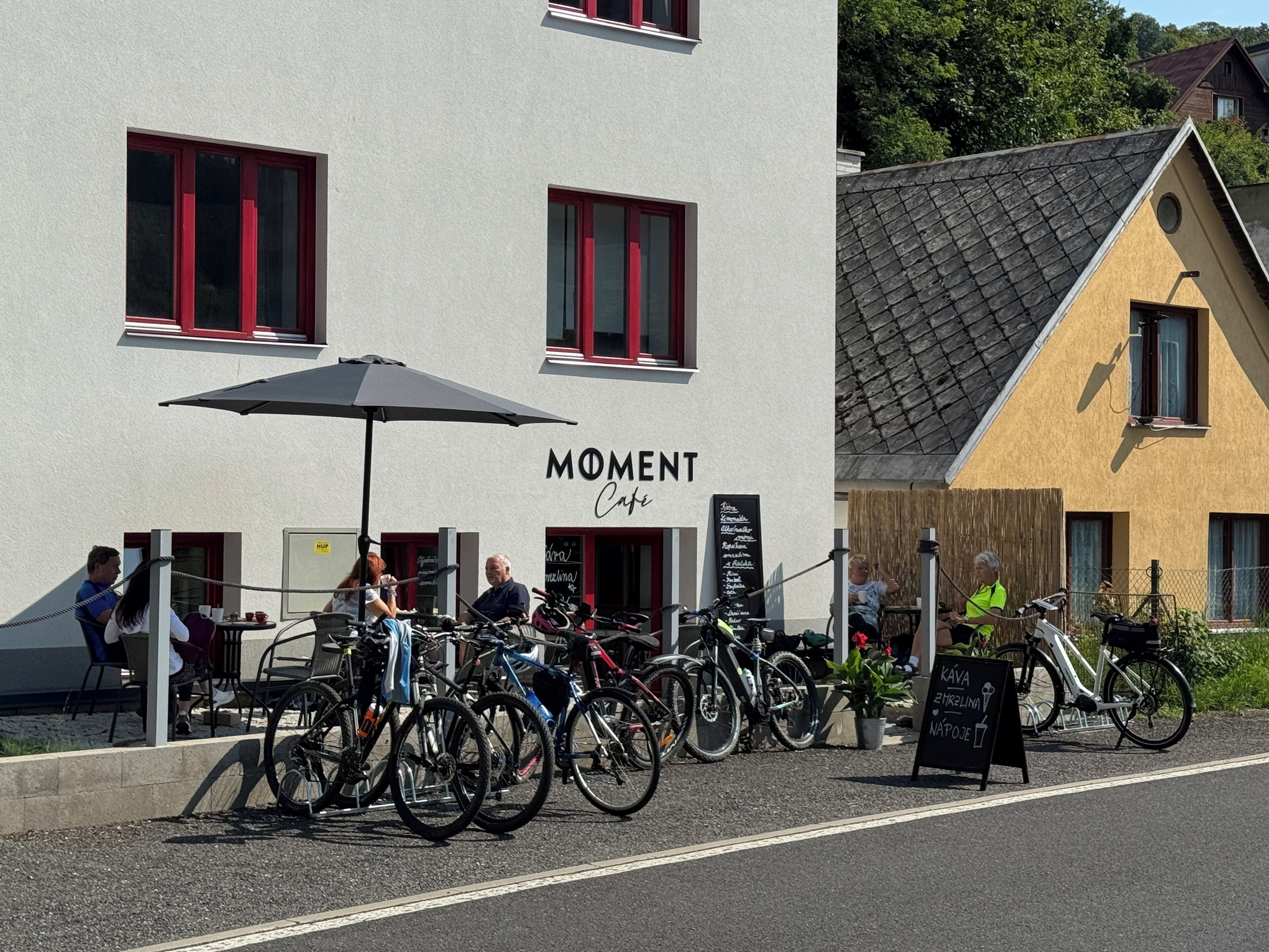 A cafe named 'Moment Cafe' with bicycles parked outside and a signboard for 'PAVA 21Klim NAPOJE'.