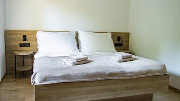 Ein ordentlich bezogenes Bett mit zwei Kissen und zwei gefalteten Handtüchern, ein kleiner runder Tisch daneben und ein hölzernes Kopfteil.