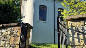 Un edificio pequeño y blanco con una sola ventana y un portón negro, rodeado de muros de piedra y vegetación.