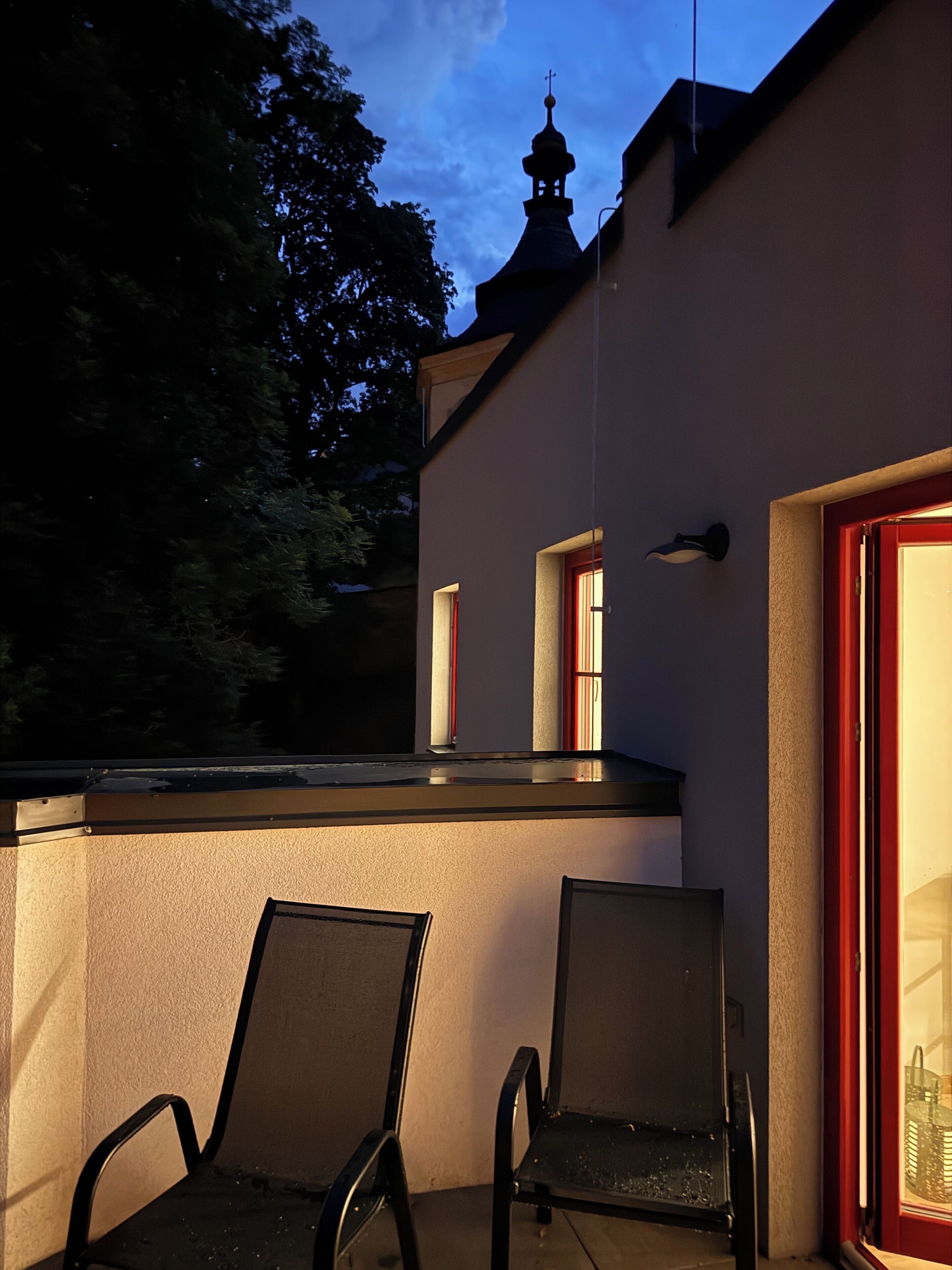Ein Sitzbereich im Freien mit zwei Stühlen und einem Tisch, ein Gebäude mit roter Tür und kleinem Fenster sowie ein Kirchturm im Hintergrund.