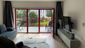 Una sala de estar moderna con un sofá, un televisor sobre un mueble bajo y un ventanal corredizo que da a un balcón exterior con sillas y barandilla roja.