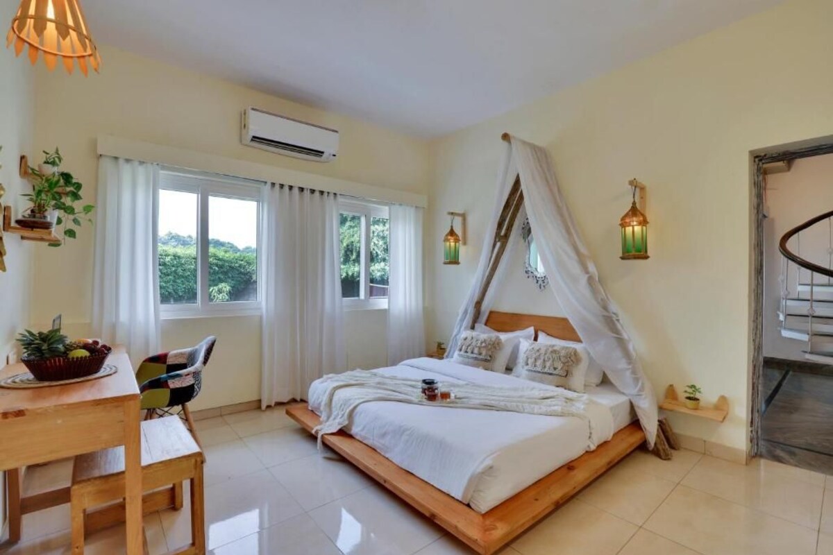 A bedroom with a wooden bed, white bedding, a wooden desk, a chair, a window with curtains, an air conditioner, and a small plant.