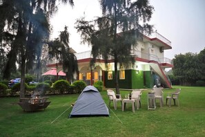A tent set up in a grassy area with a fire pit, outdoor furniture, and a building with a red and white facade in the background. - Alaya Stays By Elivaas Luxe Farm Acre - Manesar (Taoru)