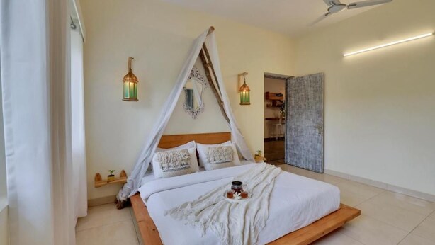 A bedroom with a canopy bed, two wall-mounted lanterns, a ceiling fan, and a door leading to another room. - Alaya Stays By Elivaas Luxe Farm Acre - Manesar (Taoru)