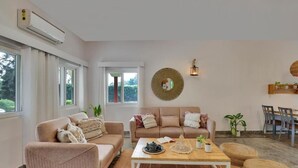 A living room with a sofa, armchair, coffee table, and a dining area with chairs and a table. - Alaya Stays By Elivaas Luxe Farm Acre - Manesar (Taoru)