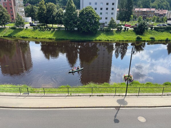 A calm river with a small boat, surrounded by greenery and buildings in the background.