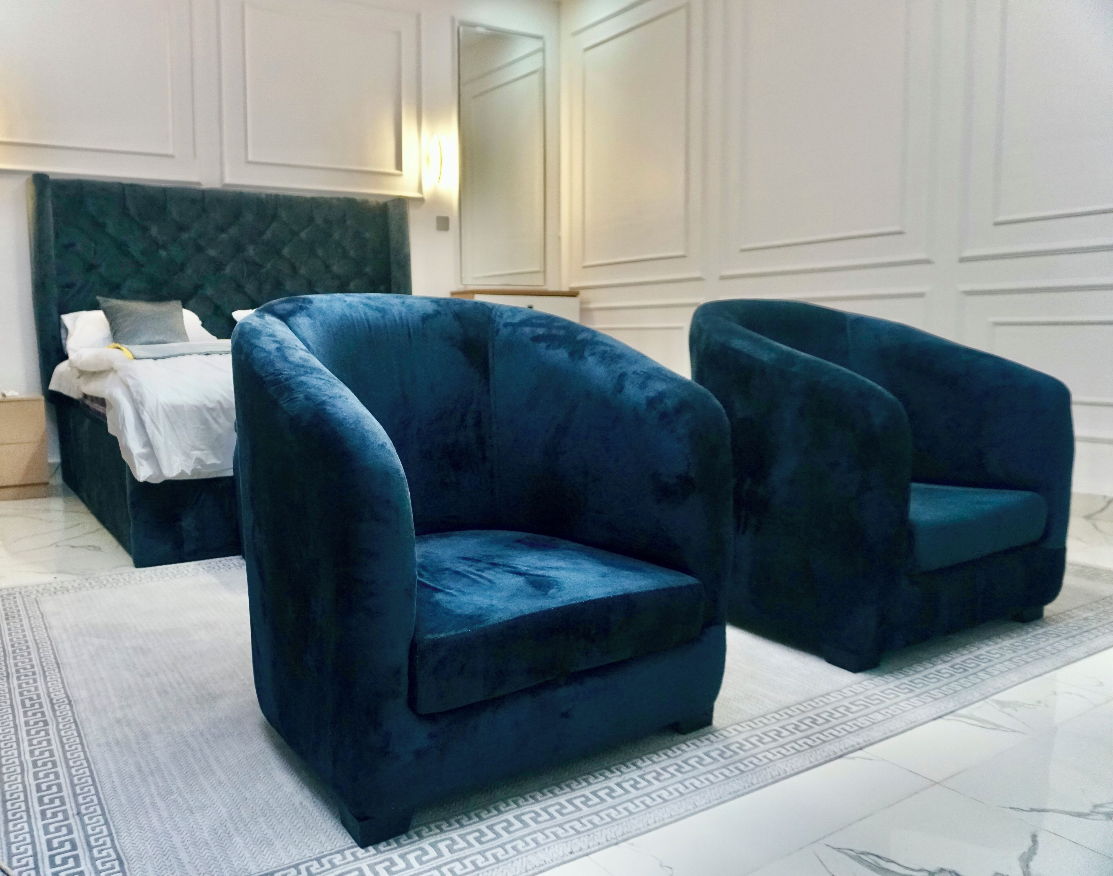 A room with a dark green upholstered armchair, a matching armchair, and a bed with a white headboard.