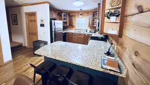 Cocina con encimeras de granito, gabinetes de madera, refrigerador de acero inoxidable y barra con taburetes.