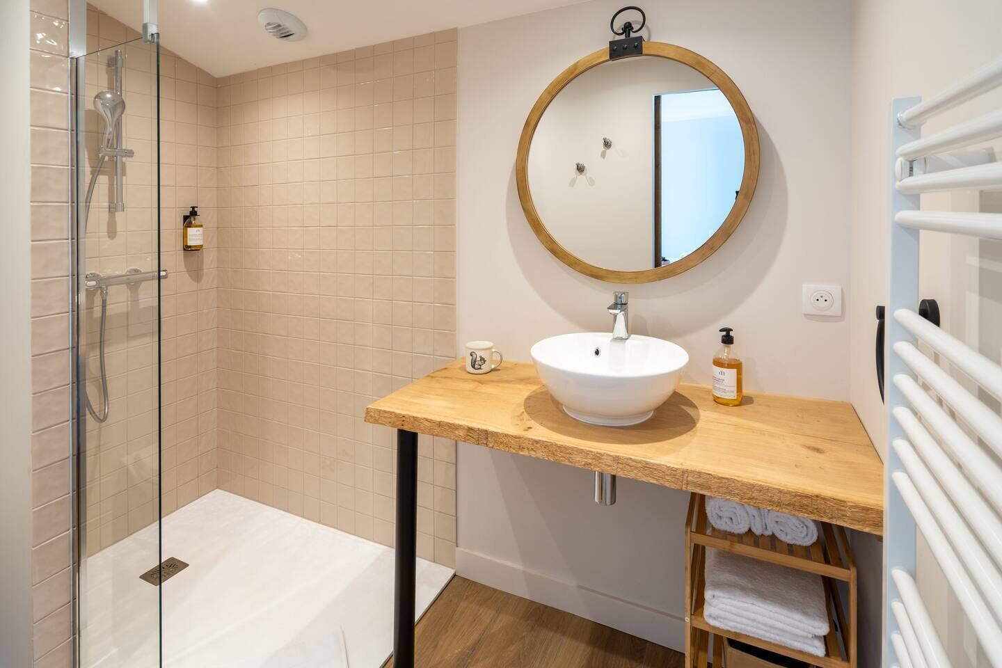 A modern bathroom with a glass shower enclosure, a wooden vanity with a white sink, a round mirror, and a towel rack.