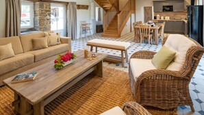 Ein großzügiges Wohnzimmer mit einer Couch, einem Holztisch und einem Korbstuhl. Eine Treppe führt zu einer Küchenzeile mit Esstisch und Stühlen.
