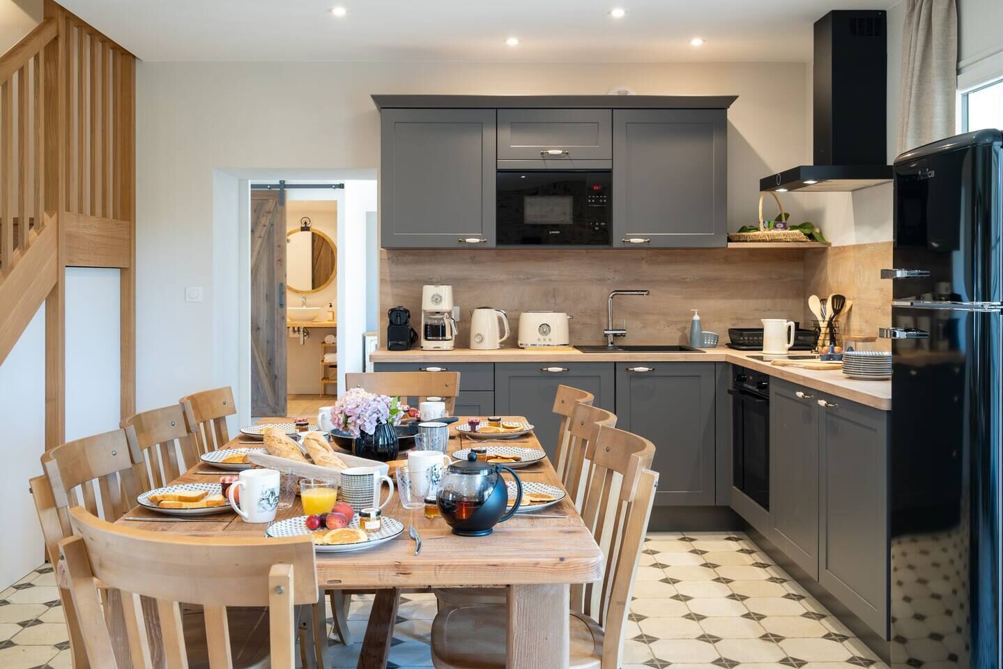 Una cocina moderna con armarios grises, un refrigerador negro y una mesa de comedor lista para el desayuno.
