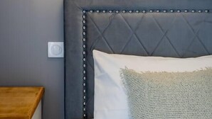 Et soveværelse med en polstret sengegavl, en vægmonteret lampe med en hornformet lysarmatur, et natbord med en træplade og en pude med struktur.