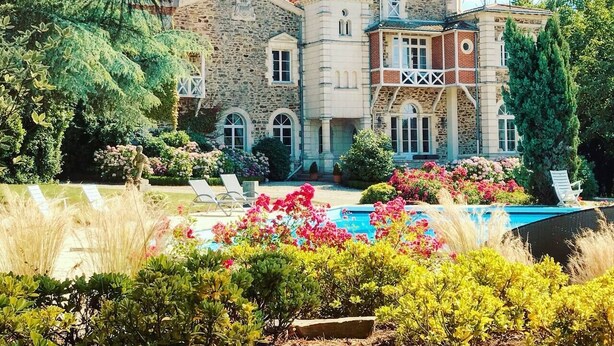 A large stone building with a swimming pool, surrounded by lush greenery and flowers.