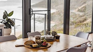 A dining table set with bread, cheese, tomatoes, and a pear, overlooking a coastal landscape. - The Nook - relaxing home breathtaking ocean views (Wye River)