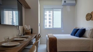 Un dormitorio con cama, ventana, una mesa de comedor con sillas y televisor montado en la pared.