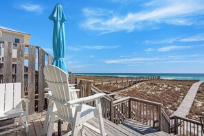 A wooden deck with white chairs and a blue umbrella, overlooking a beach and ocean.