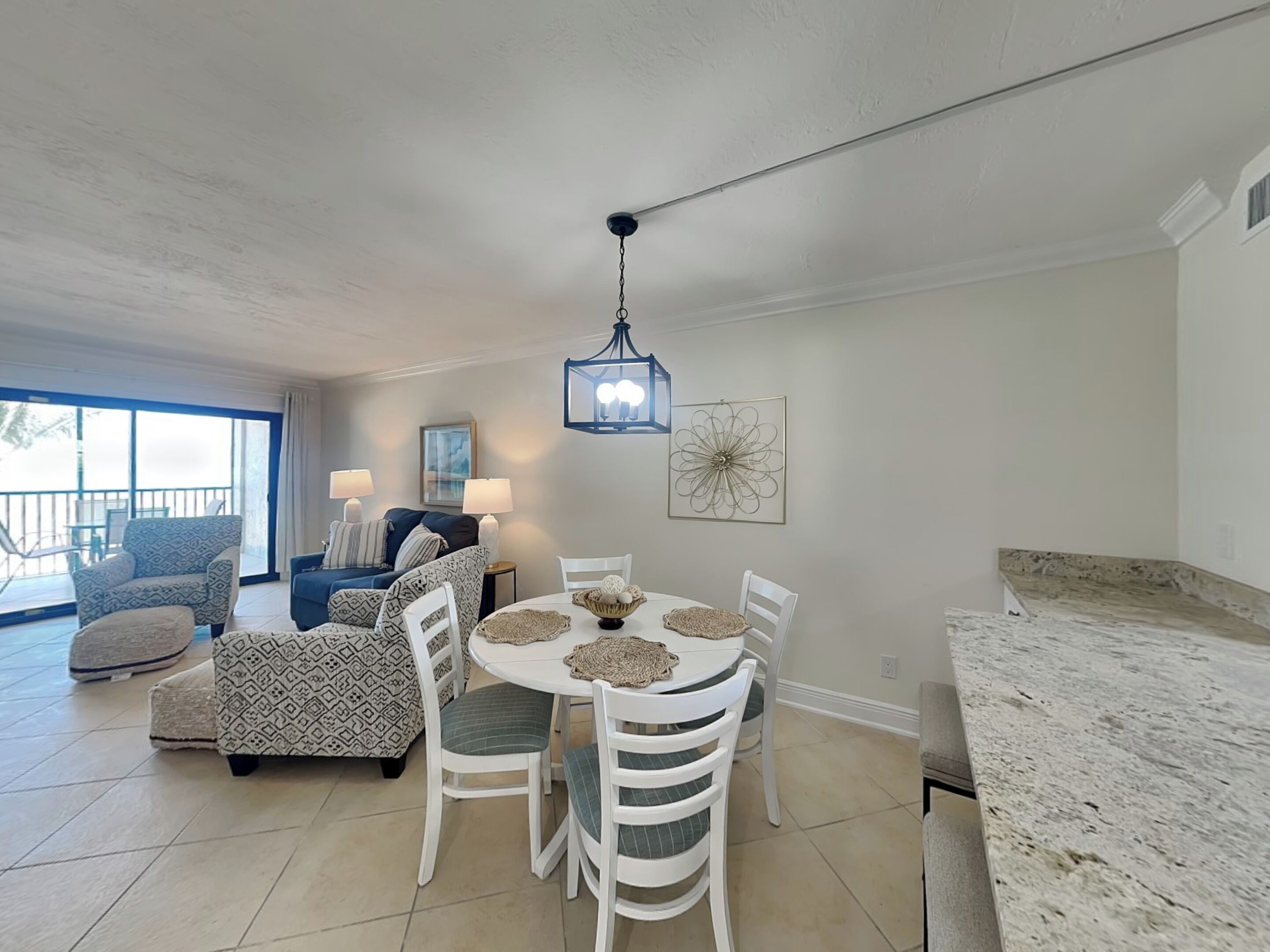 A dining area with a round table, white chairs, and a chandelier. There is a sofa and a chair with patterned upholstery in the background.