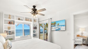 Un dormitorio con una cama grande, ventilador de techo, estantería, televisor y vista al océano.