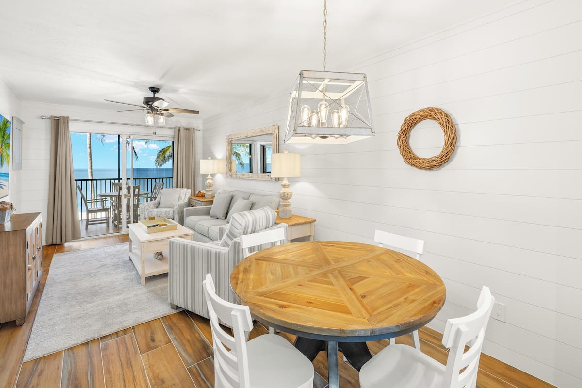 Amplia sala de estar con mesa de comedor de madera, sillas blancas, sofá y balcón con vista al océano.