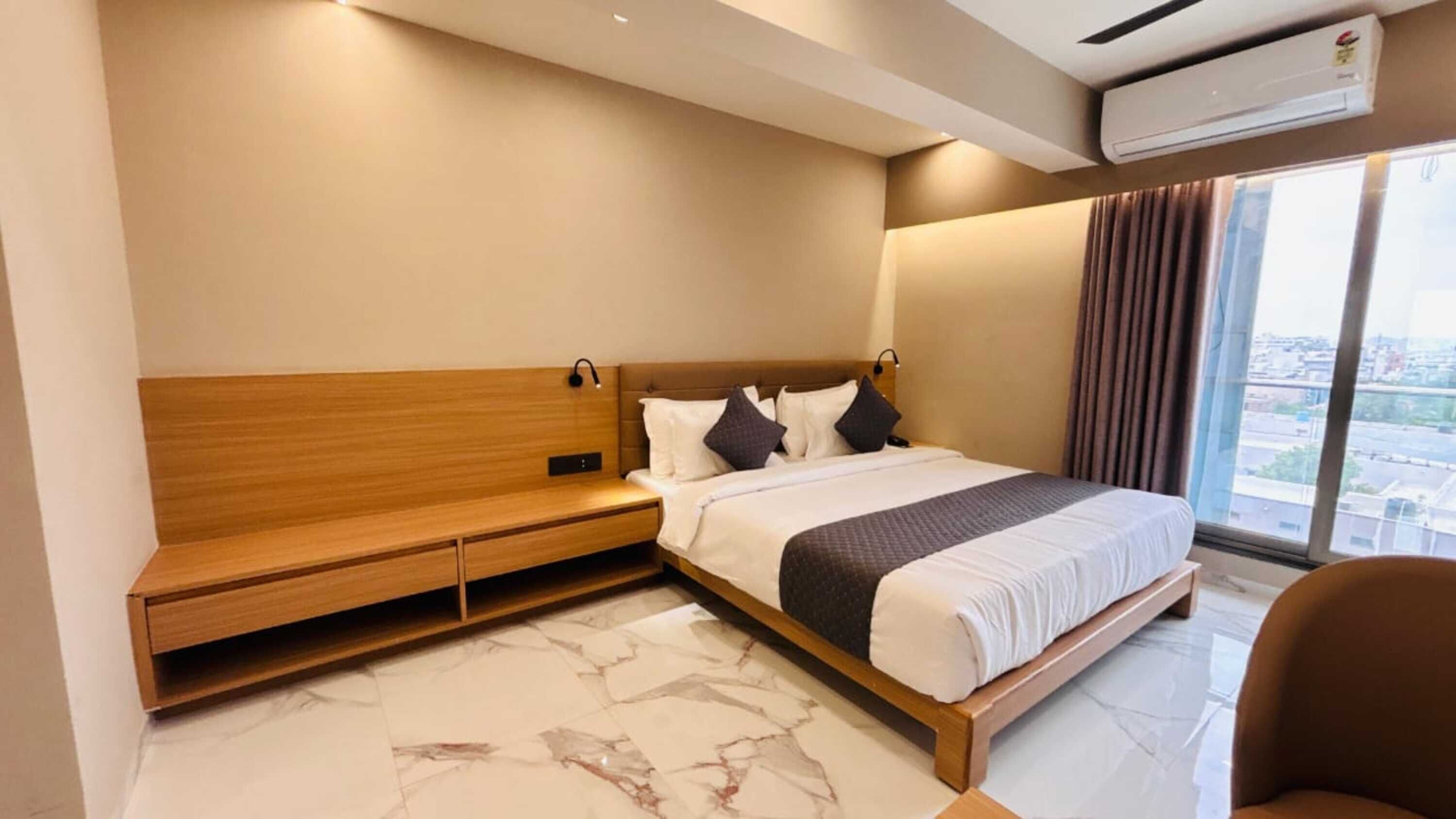 Habitación de hotel moderna con una cama grande, un banco de madera y vistas a la ciudad a través de amplios ventanales.