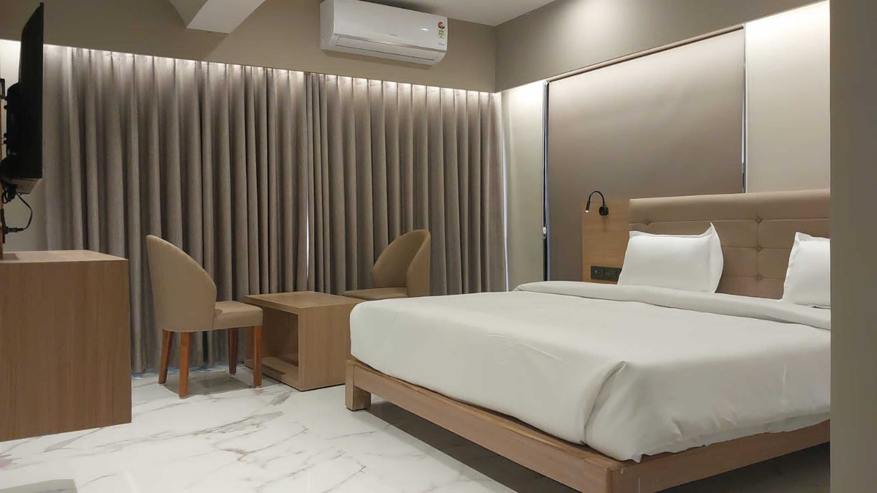 A modern hotel room with a large bed, a desk, two chairs, a TV, and a wardrobe.