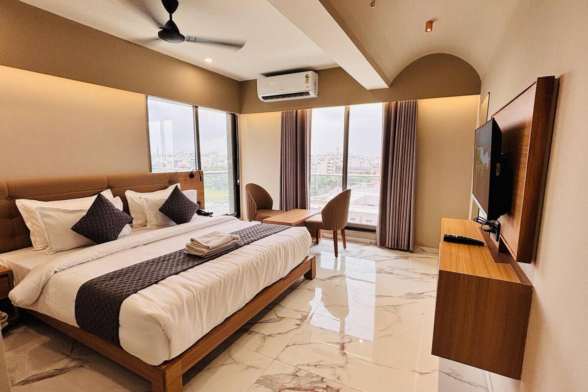 A hotel room with a large bed, a desk, a chair, a TV, and a balcony with curtains.