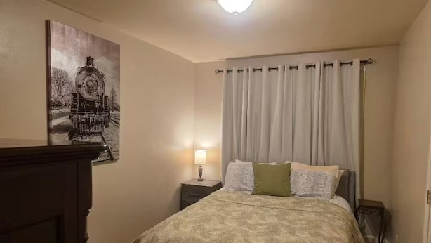 A bedroom with a bed, a nightstand, a lamp, a painting, and a window with curtains. - Home Away from Home (Chattanooga)