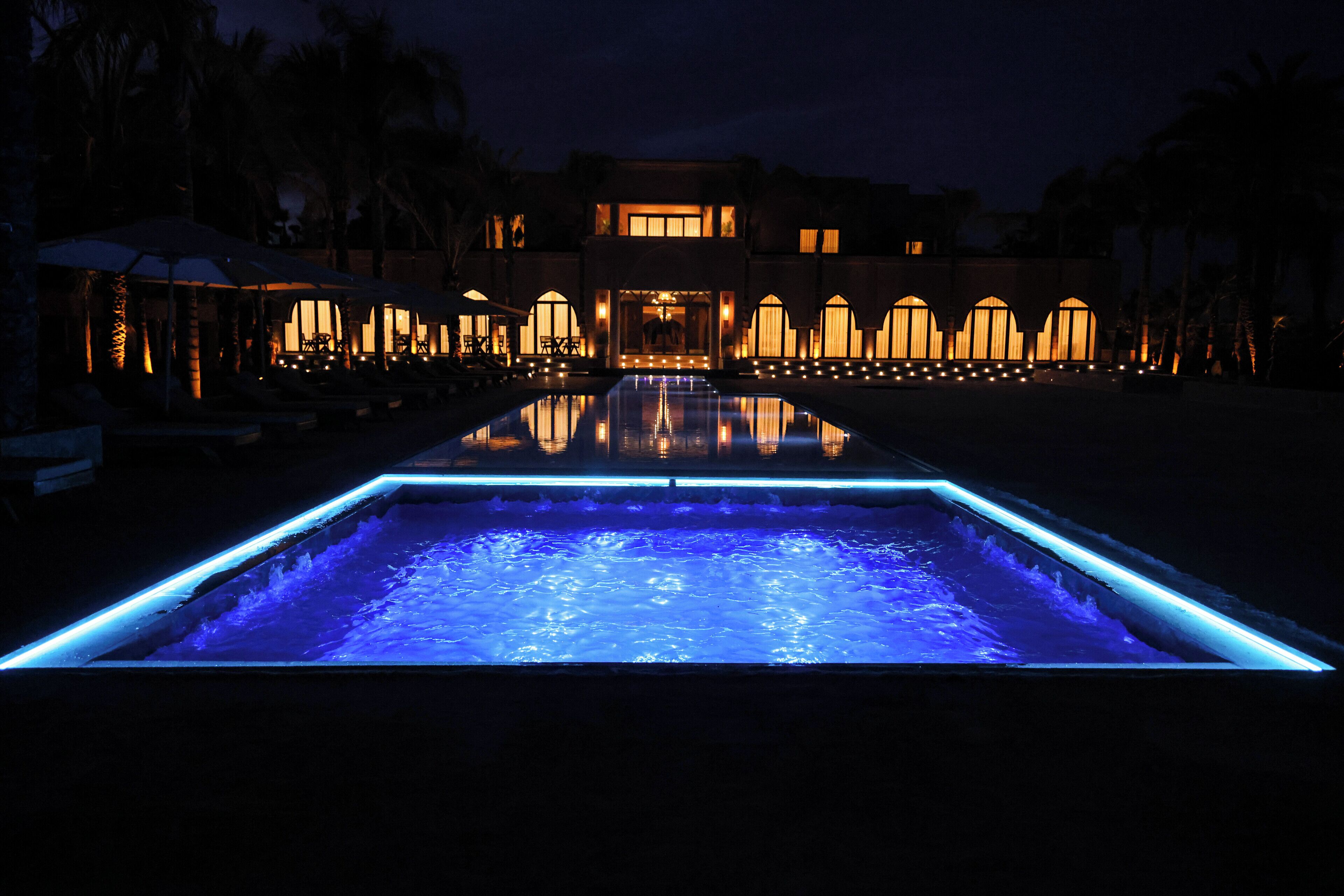 A swimming pool with a blue-lit edge, a building with arched windows, and palm trees in the background.