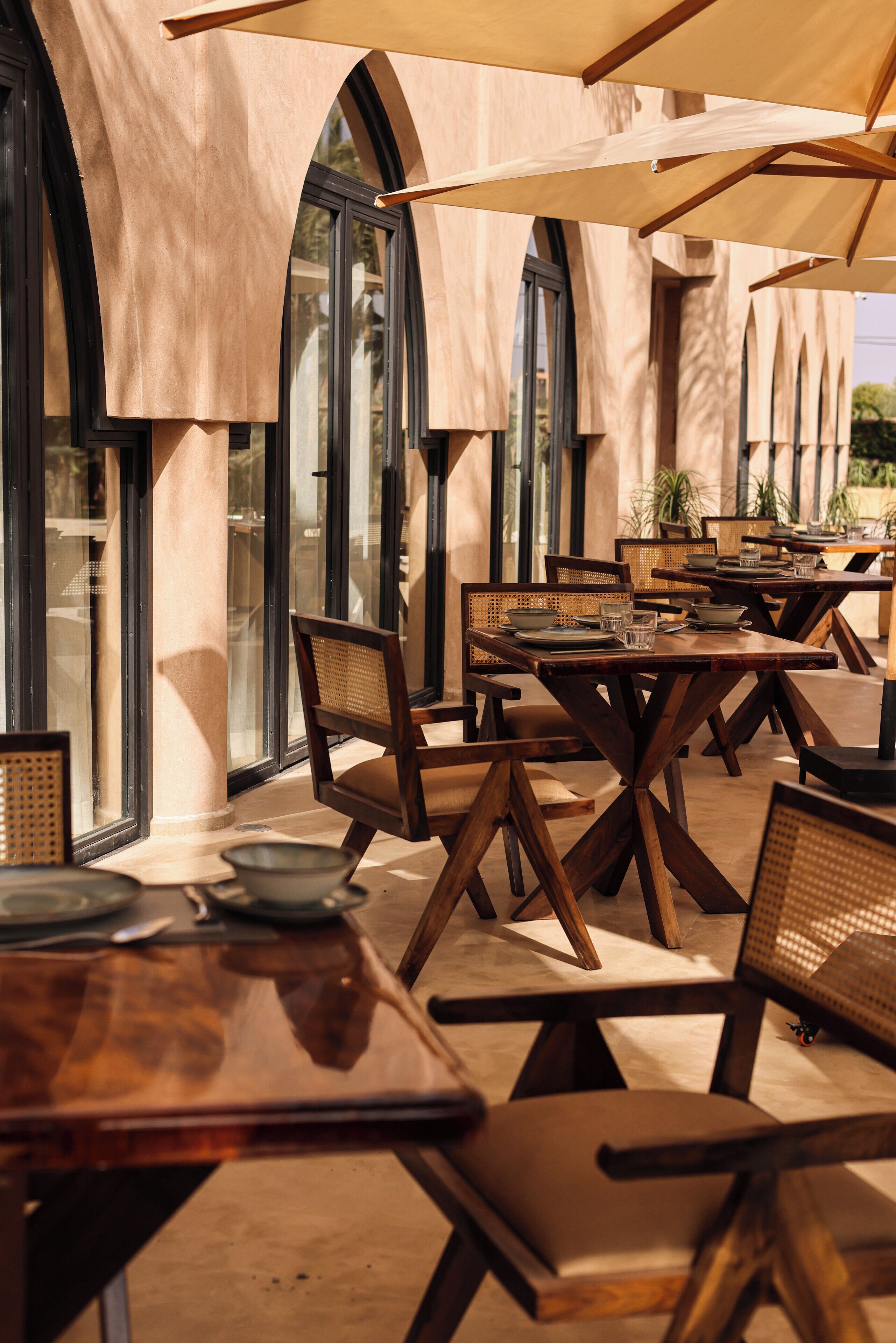 Un espace de restauration en plein air avec des tables et des chaises en bois, de grandes fenêtres et une pergola offrant de l’ombre.