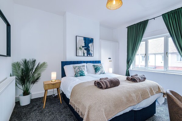 Un dormitorio con cama, mesita de noche con lámpara, planta en maceta y ventana con cortinas.