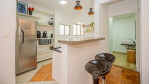 A modern kitchen with a stainless steel refrigerator, a coffee maker, and a microwave. There are two bar stools and pendant lights above the counter. - Apartamento Aconchegante a 850m da Av Paulista (São Paulo)