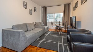 A modern living room with a grey sofa, a black leather chair, a dining table with chairs, a flat-screen TV, and a large window with curtains. - Apartamento Aconchegante a 850m da Av Paulista (São Paulo)
