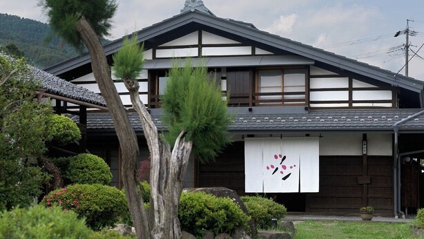 Una casa tradicional japonesa con tejado de tejas, vigas de madera y un jardín con diversas plantas y un árbol grande.