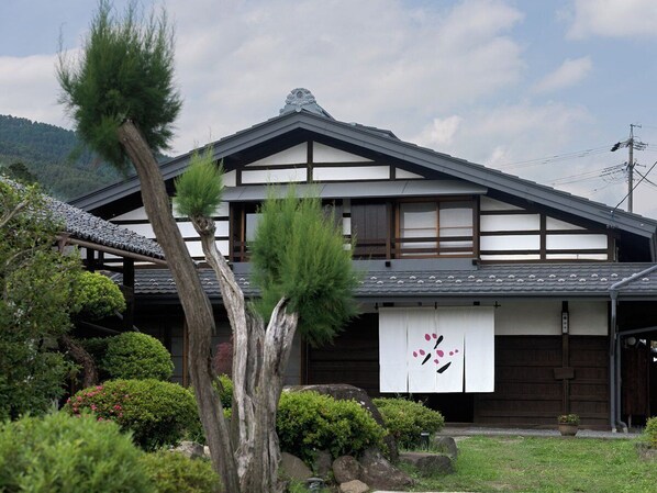 Una casa tradicional japonesa con tejado de tejas, vigas de madera y un jardín con diversas plantas y un árbol grande.