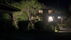 Una casa tradicional japonesa de noche, con una entrada bien iluminada y vegetación circundante.