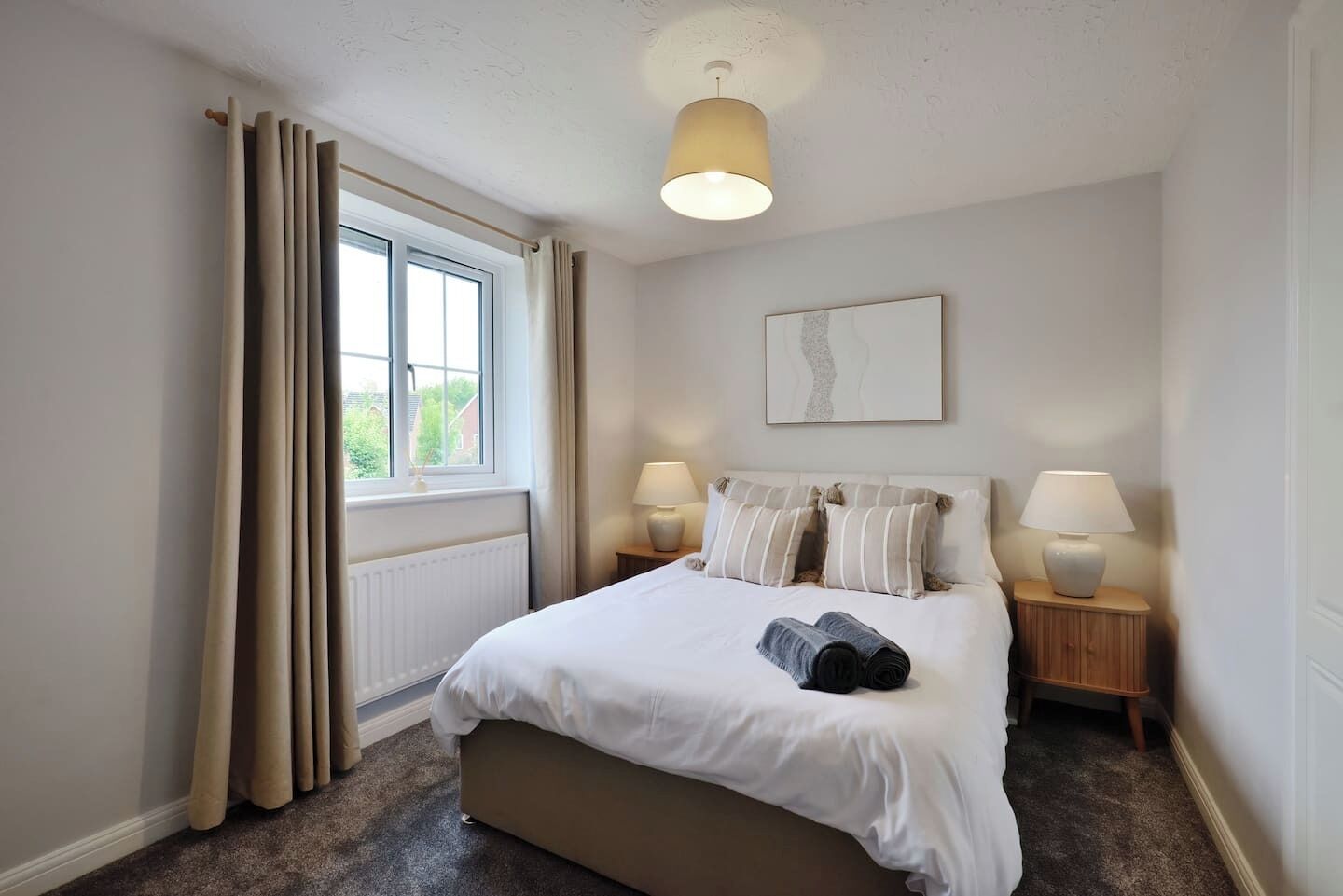 Une chambre à coucher avec un grand lit, deux tables de chevet avec des lampes, une fenêtre avec des rideaux, un radiateur et une œuvre d’art accrochée au mur.