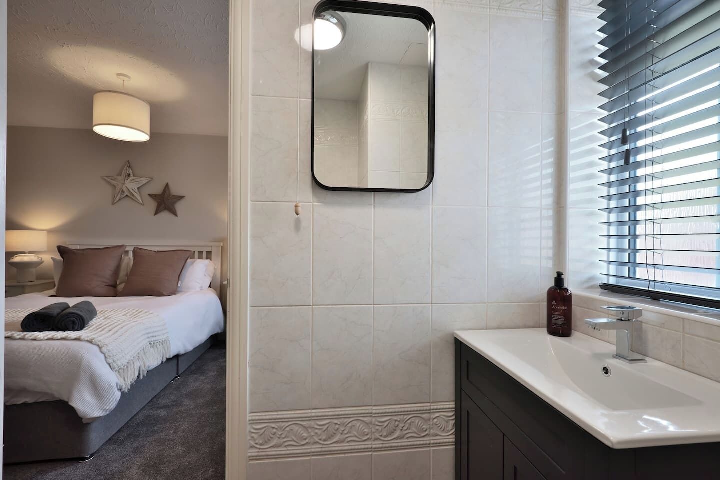 Ein Schlafzimmer mit Bett, Nachttisch und Ablagefläche. Es gibt ein Badezimmer mit Waschbecken, Spiegel und einem Fenster mit Jalousien.