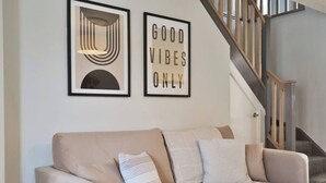 A living room with a beige sofa, two framed artworks, and a staircase.